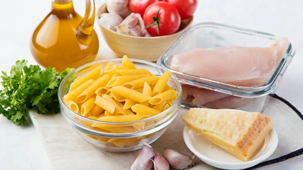 A collection of ingredients for garlic Parmesan chicken pasta, including fresh chicken breasts, garlic cloves, grated Parmesan cheese, pasta, heavy cream, and fresh herbs.